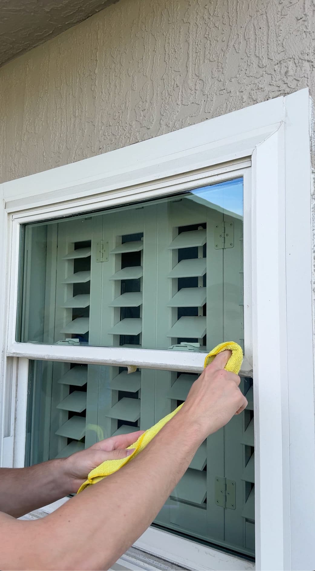 Person cleaning a window with a yellow cloth, revealing a green shutter background.