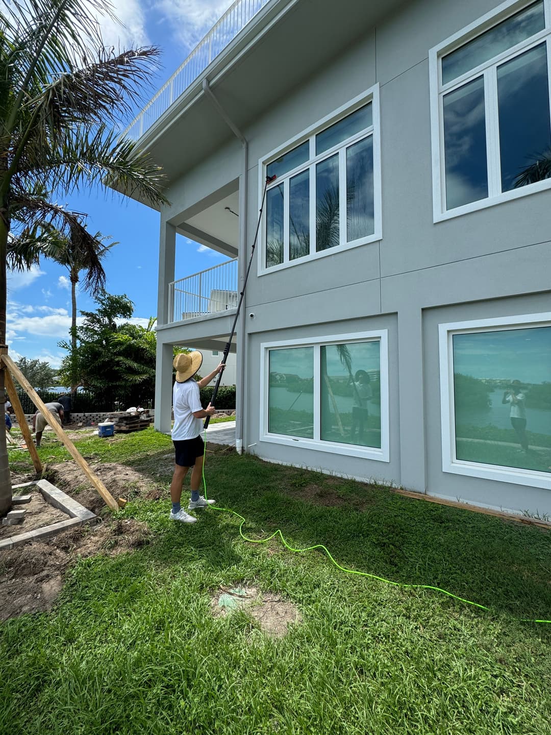 Person painting a house exterior with a roller on a pole, surrounded by greenery and palm trees.