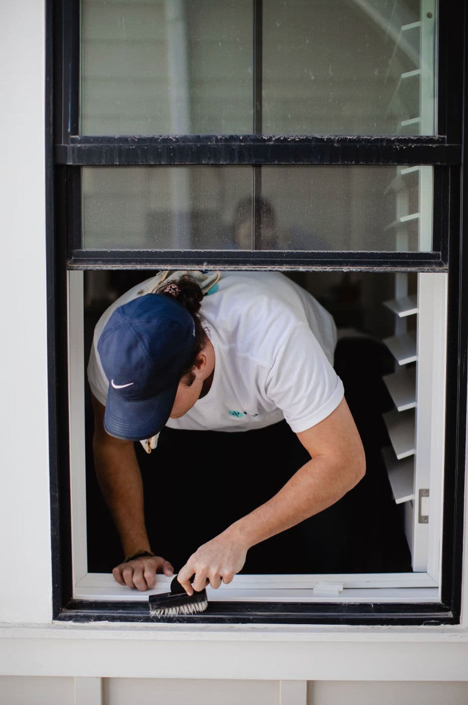 Person cleaning a window from inside a house, wearing a cap and a white shirt.