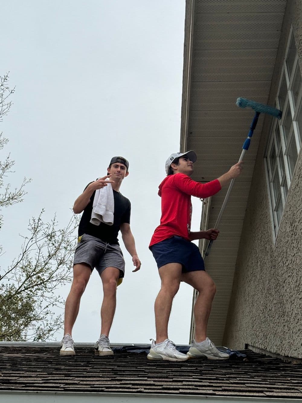 Two young men cleaning windows from a roof, using a mop and towel, on a cloudy day.