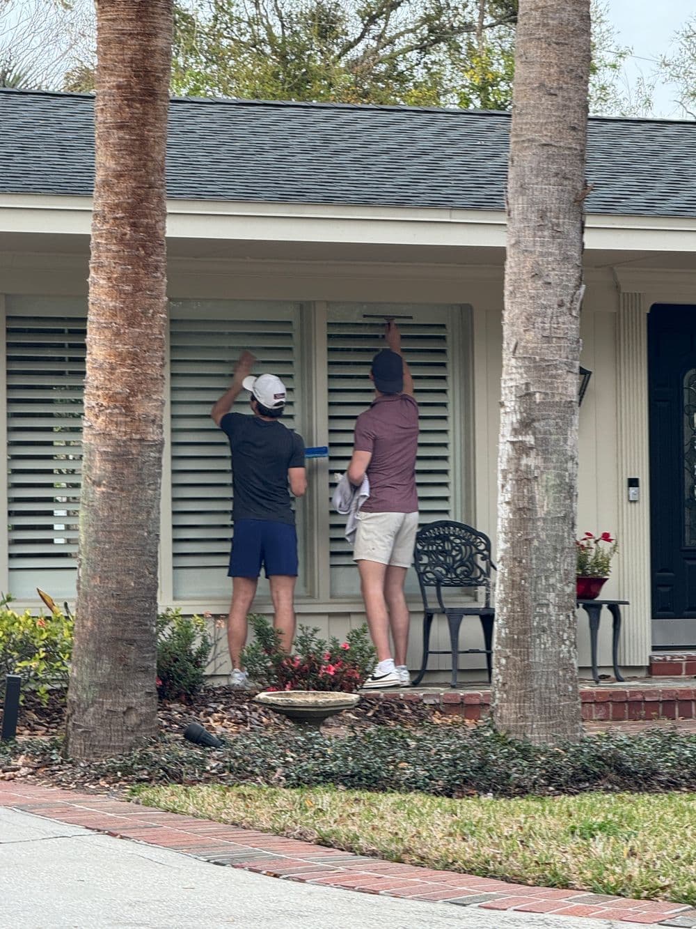 Two men cleaning window shutters on a house surrounded by palm trees and flowers.