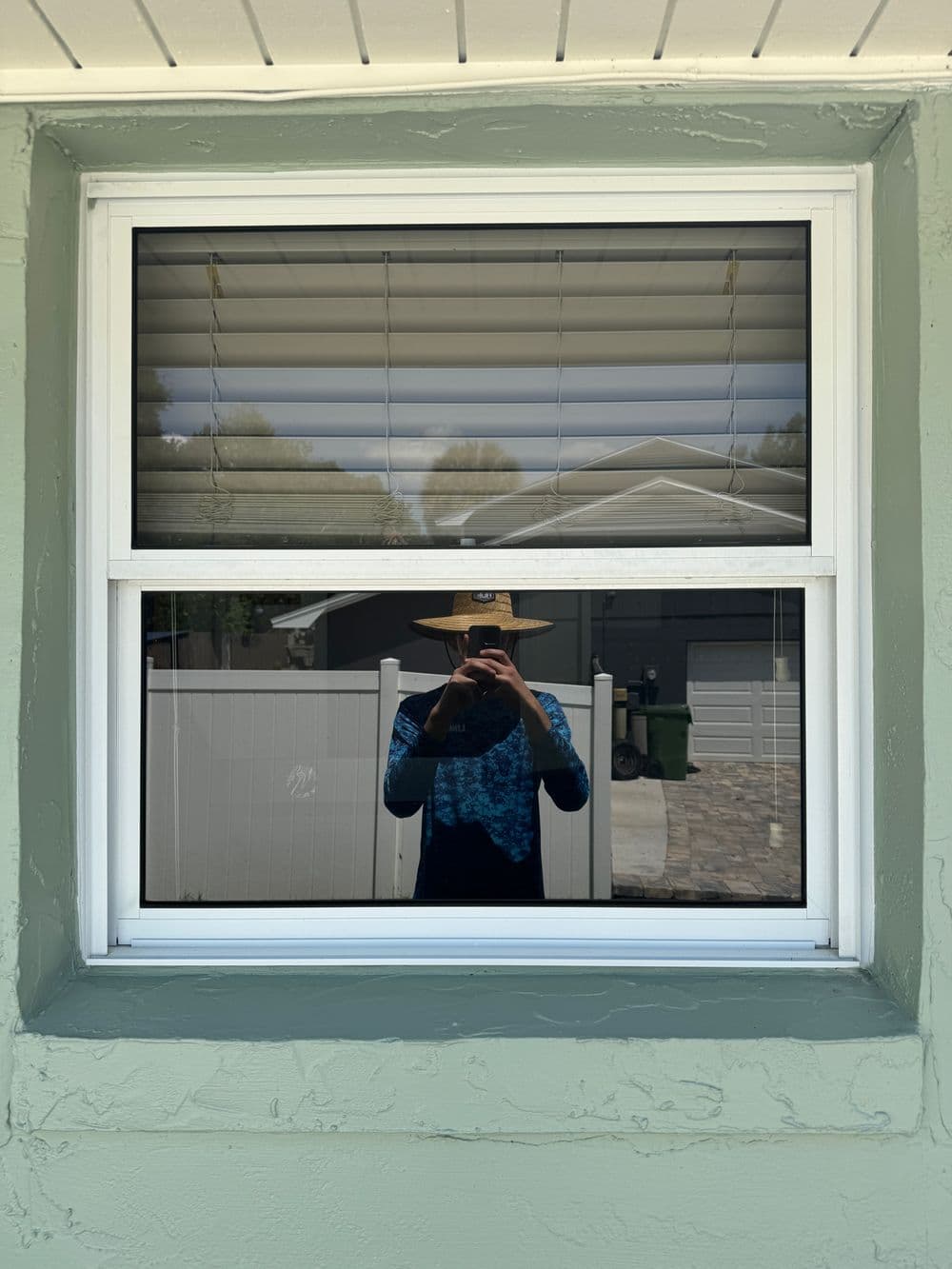 Reflection of a person in window with blinds, wearing a hat, standing outside a house.