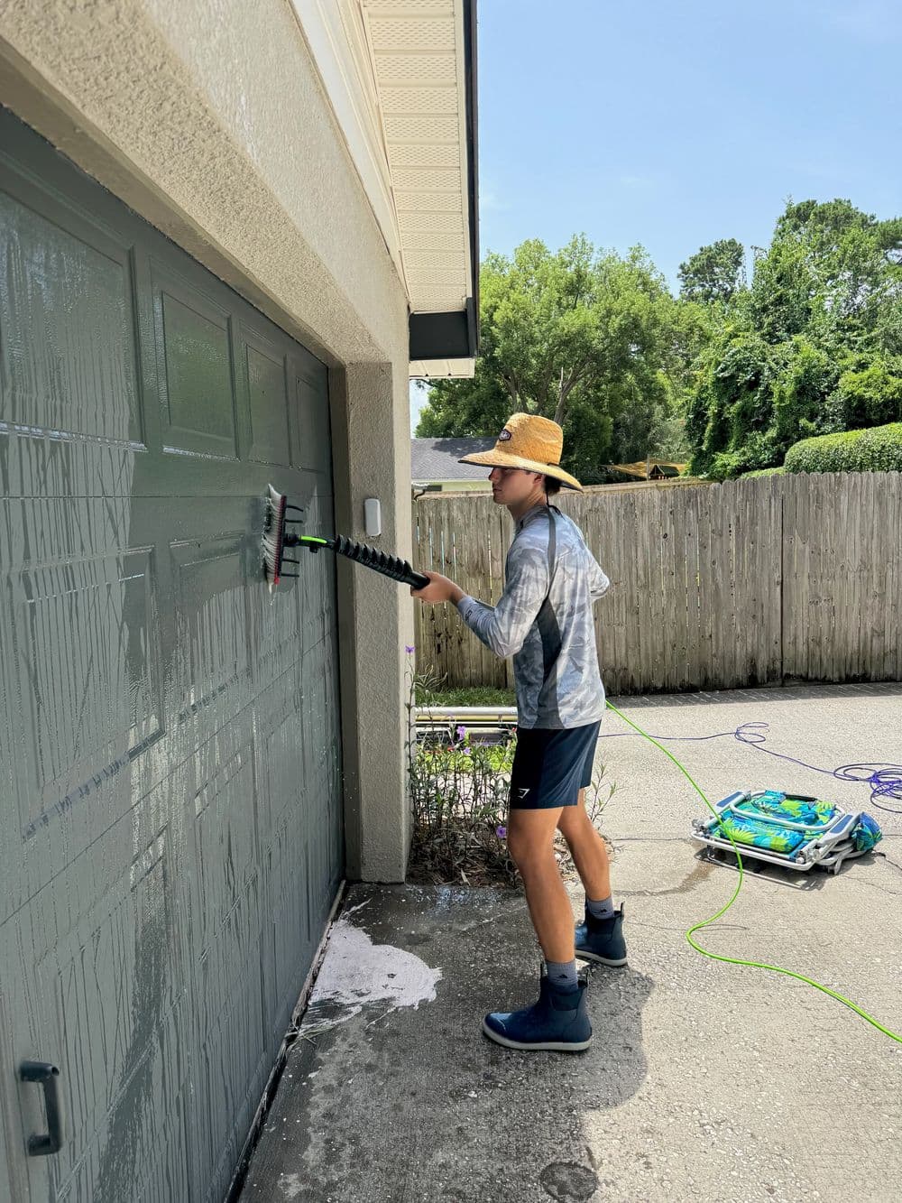 Person wearing a straw hat cleaning a garage door with a long brush in sunny weather.