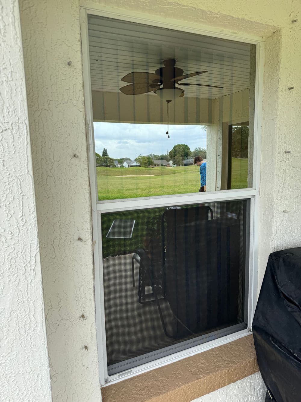 View through a screened window showing a porch chair and a person outdoors.