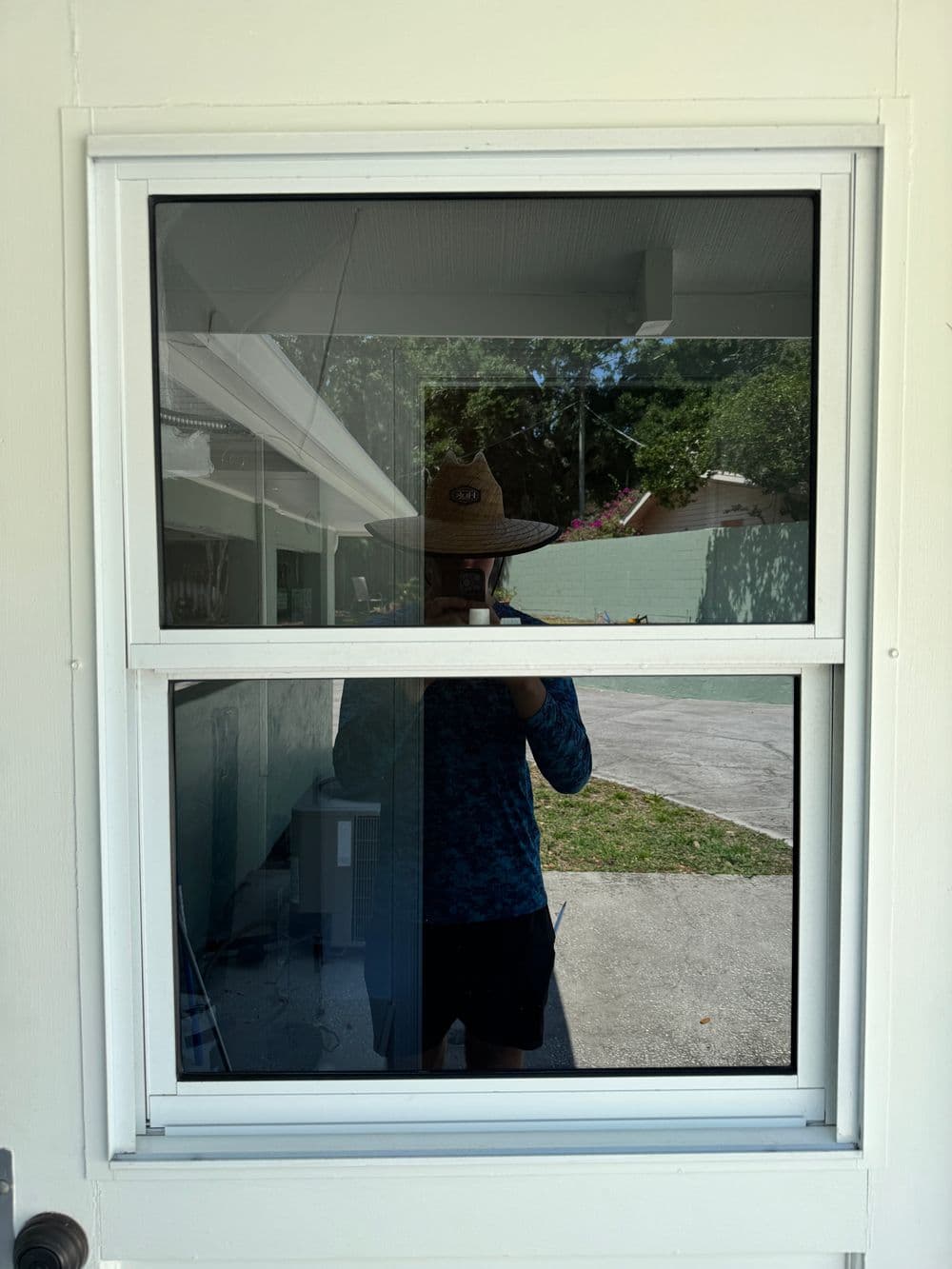 Person wearing a sun hat reflected in a window, with a garden and house visible outside.