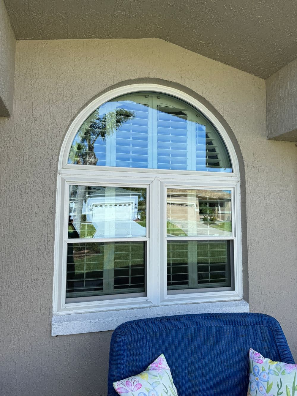 Arched window with shutters and a blue chair on a porch with colorful cushions.