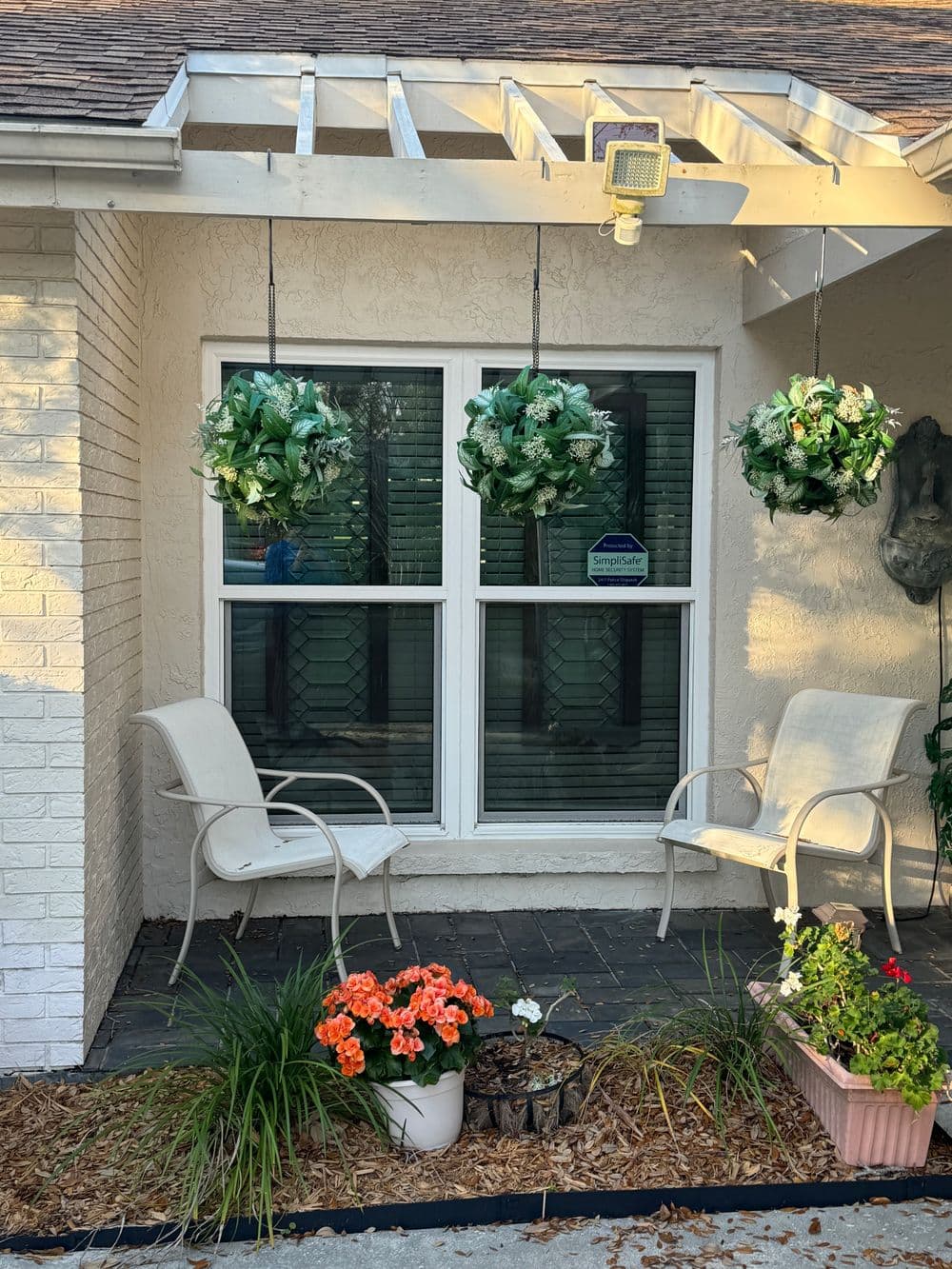 Front porch with hanging greenery, two chairs, and colorful flower pots by the window.