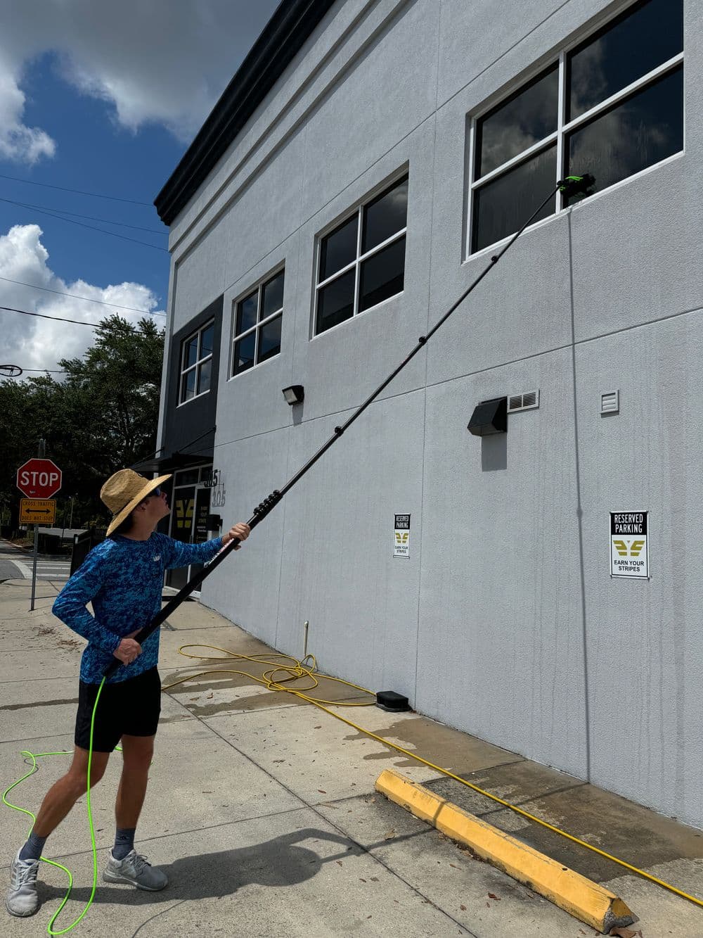 Person using a high-pressure washer on a building window with a long pole, sunny day.