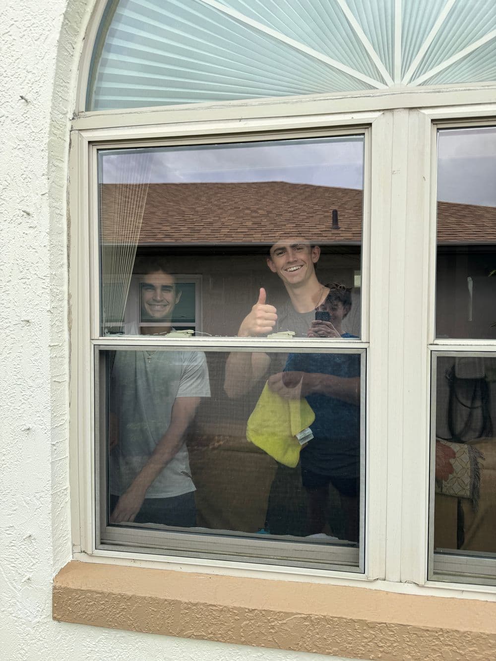 Two young men smiling and giving a thumbs up from inside a house window.