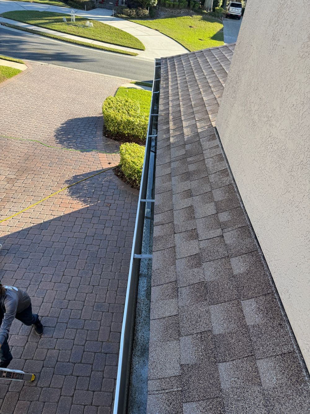 Roof gutter cleaning service with a worker on a ladder near a paved driveway.